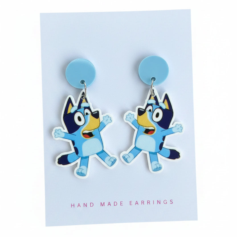A pair of cartoon dog stud earrings displayed on a white backing card. The earrings feature a light blue circular stud and a dangling die-cut character charm in shades of blue, yellow, and black, with a small gold-tone ring at the top.